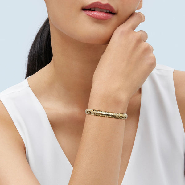 Model in a white top, hand at the chin, wearing a ribbed gold bracelet